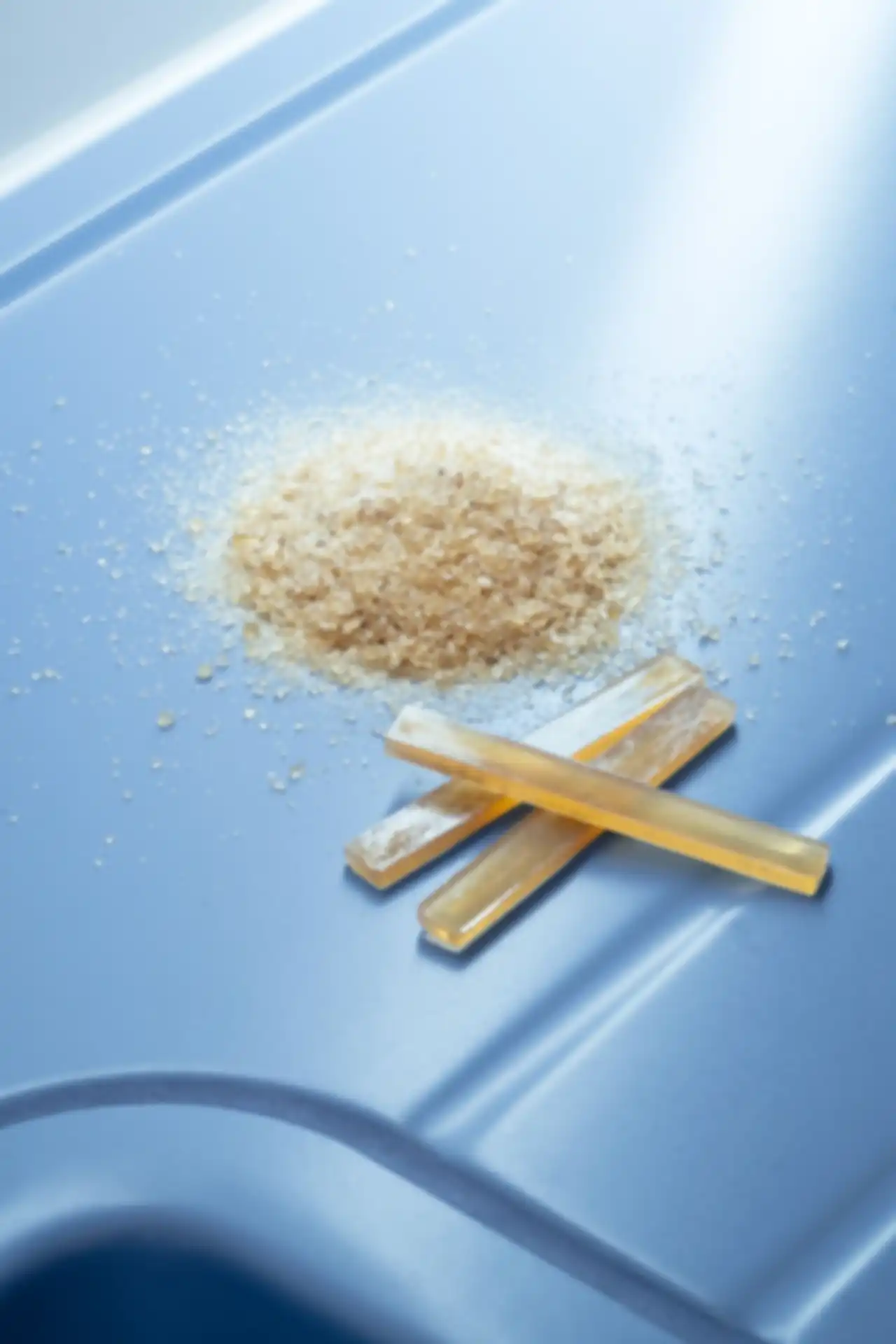 Ein leicht gelbliches, pulverartiges Material liegt neben drei plastikähnlichen Sticks auf einem neutralen Grund