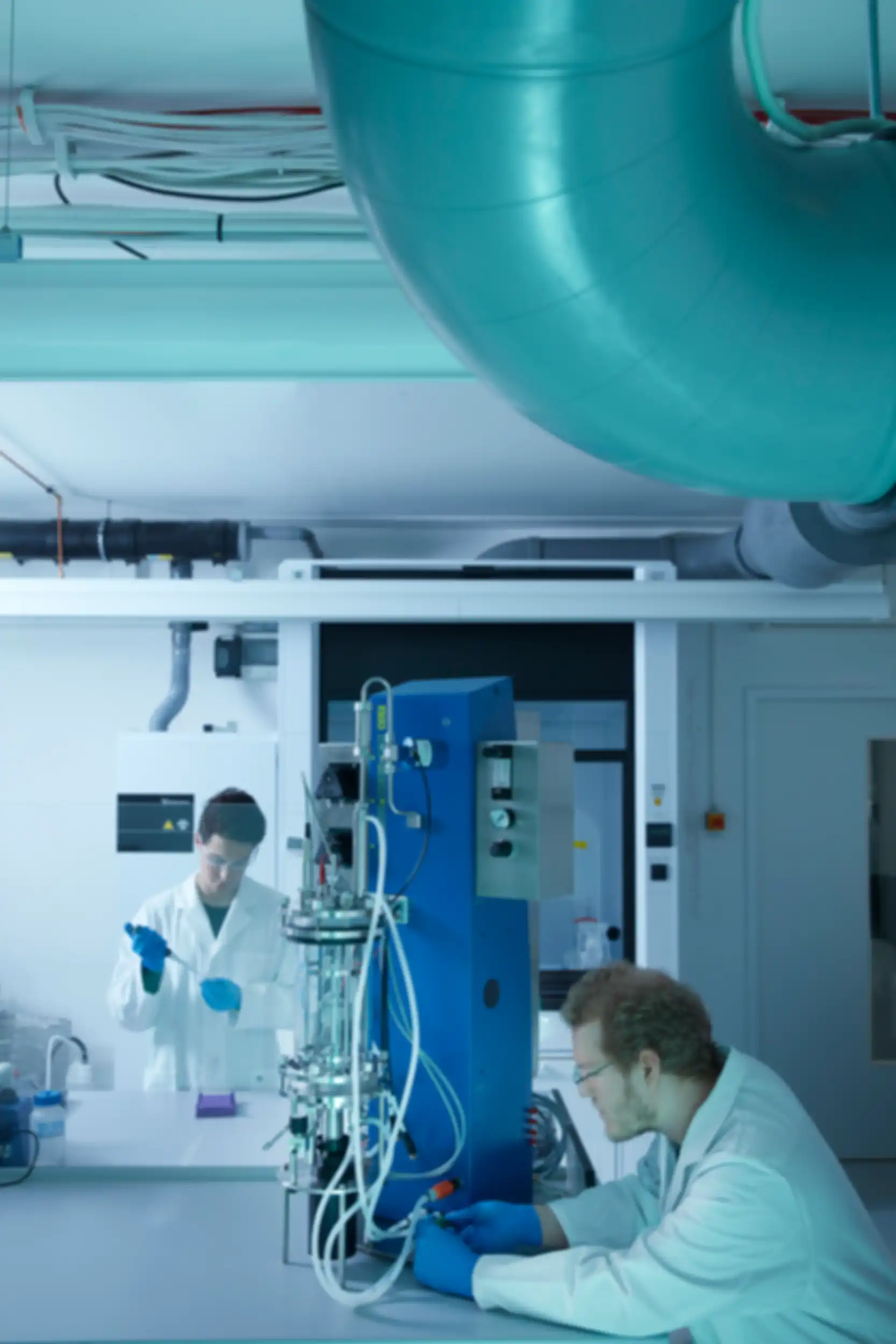 A man is operating a piece of technical equipment. Behind a glass pane, another man is holding a pipette.