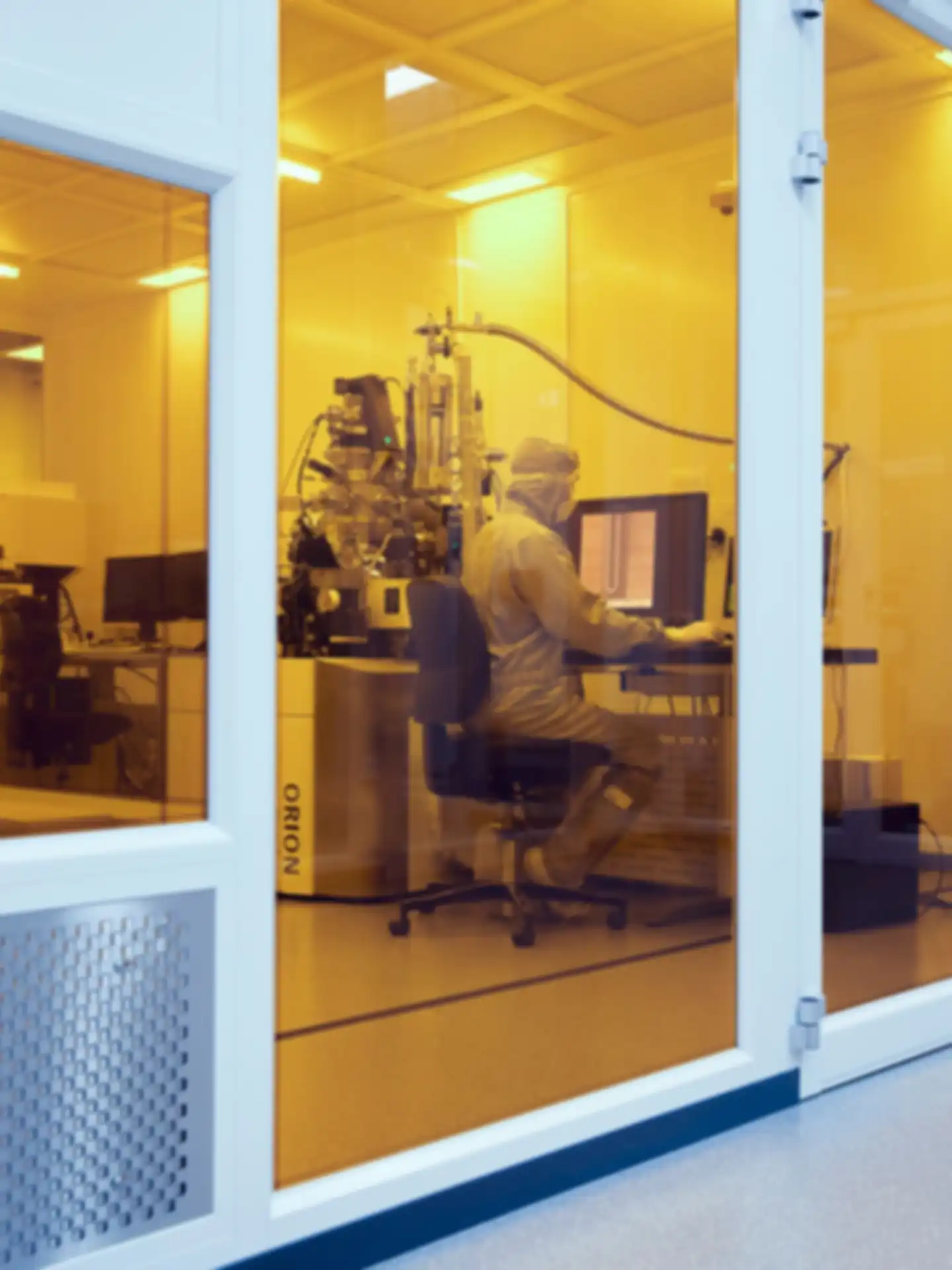 Eine Person mit Ganzkörperanzug und Brille sitzt an einem Computer in einem Labor, umgeben von technischen Apparaturen