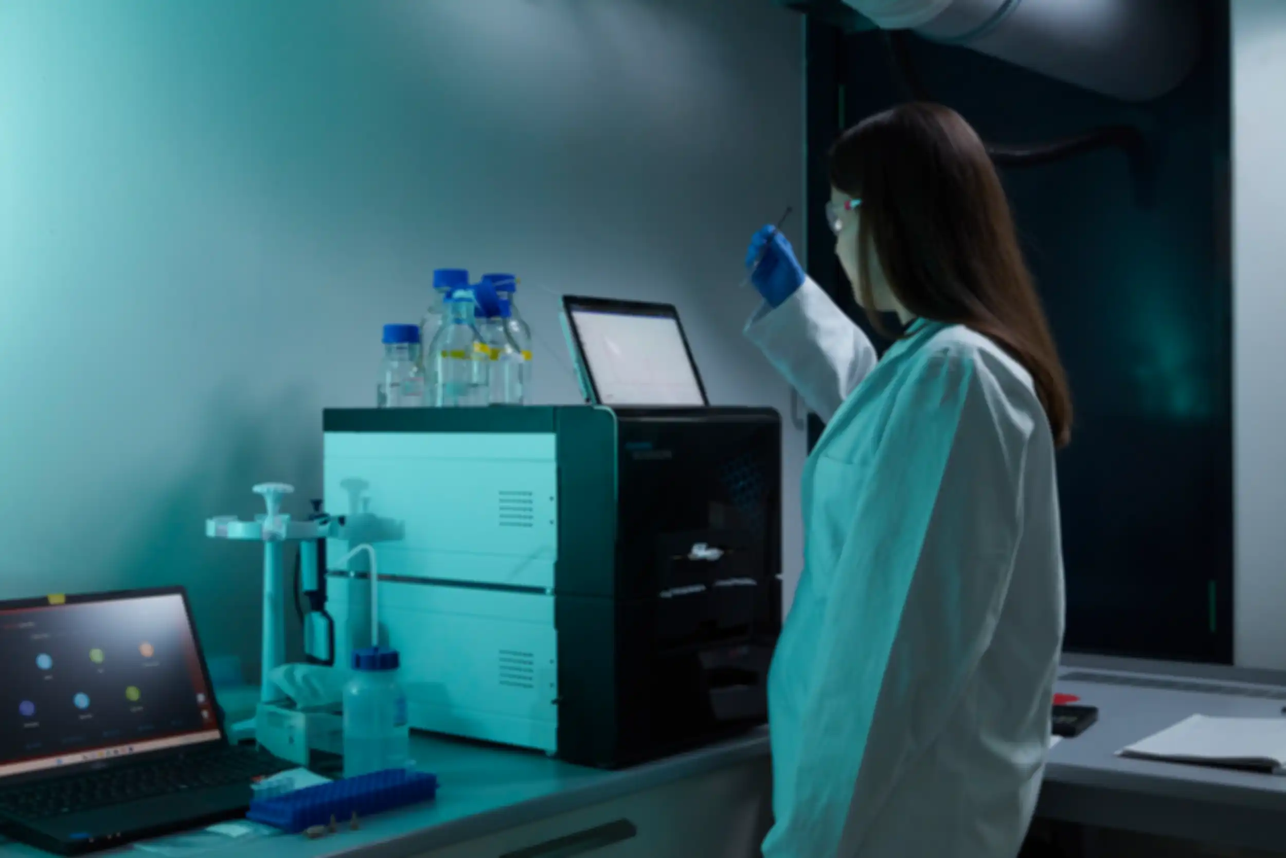 A woman wearing safety goggles and a lab coat is examining a sample in front of a laptop displaying a graph