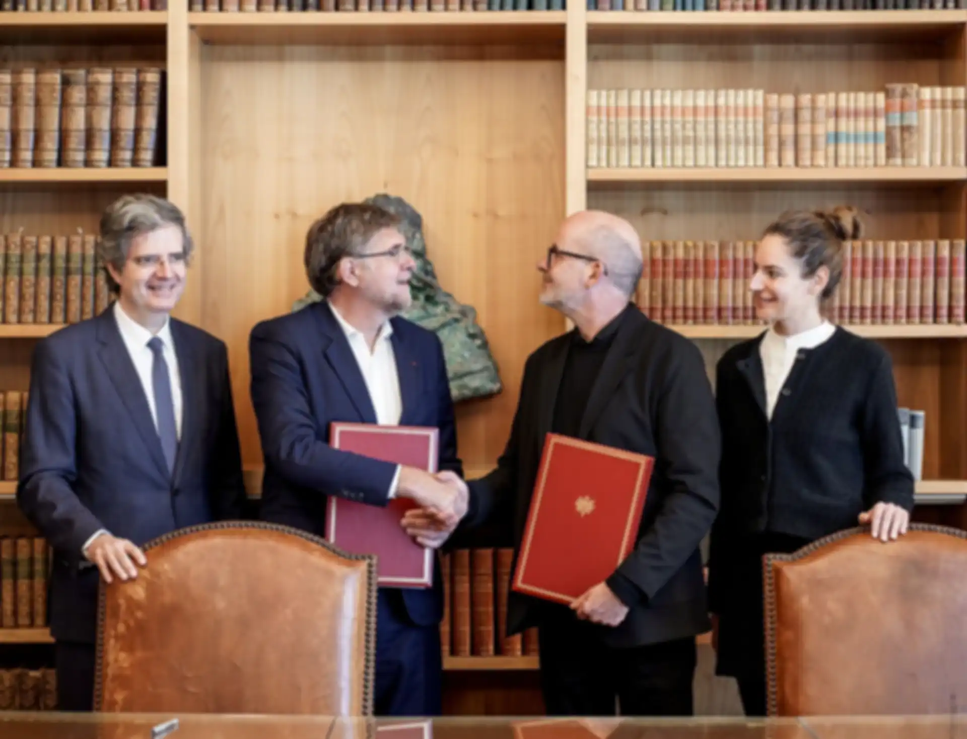 Two people shake hands after signing the agreement. Two people are standing nearby