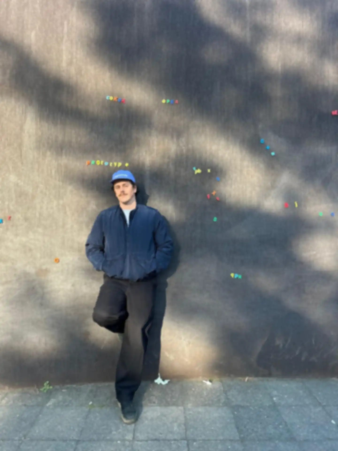 A full-body portrait of Andreas Klinger, leaning with his back against a gray wall covered with colorful letters. One leg is bent, and both hands are in his jacket pockets.
