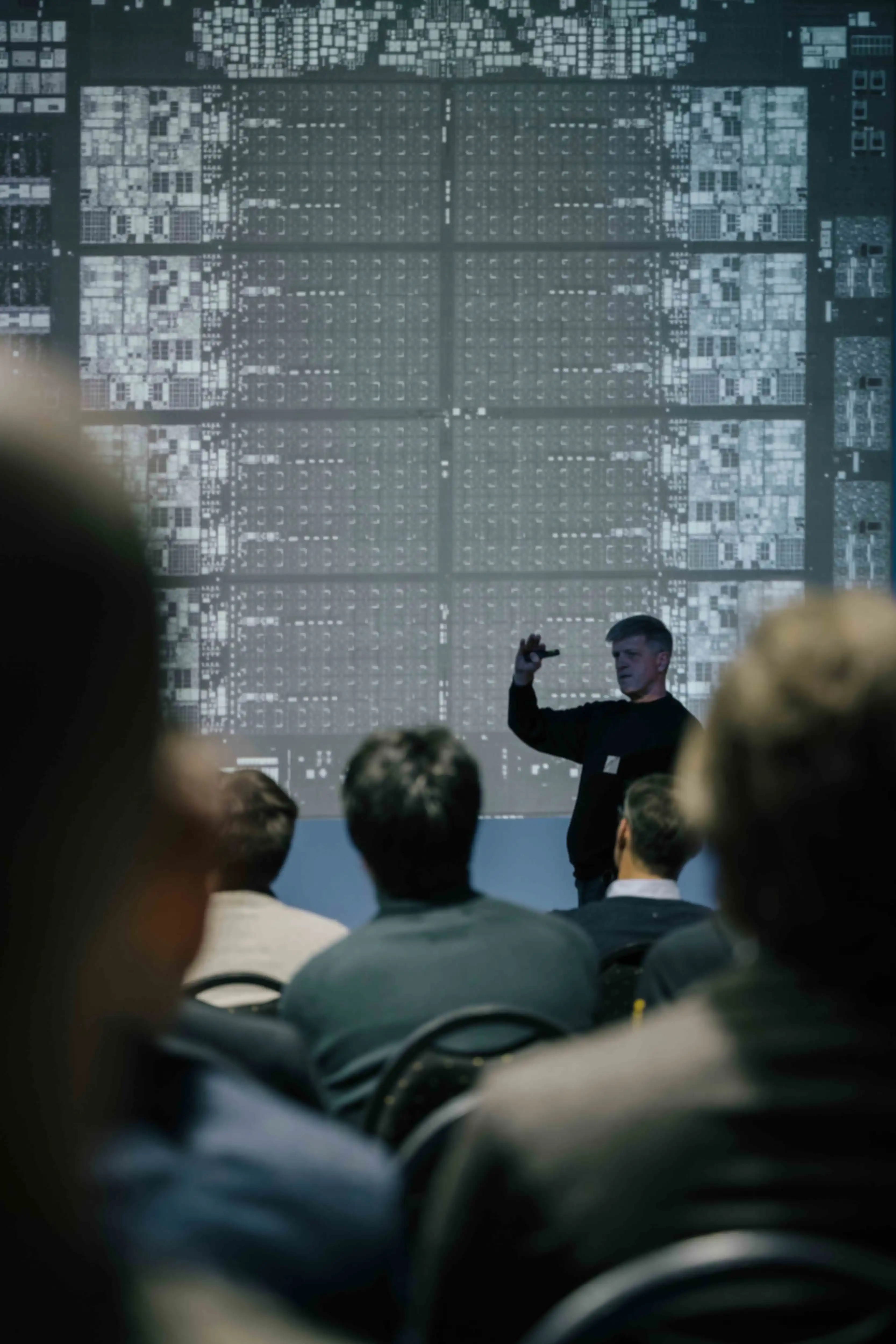 A person is giving a presentation. In the background is a large screen displaying a black-and-white graphic.
