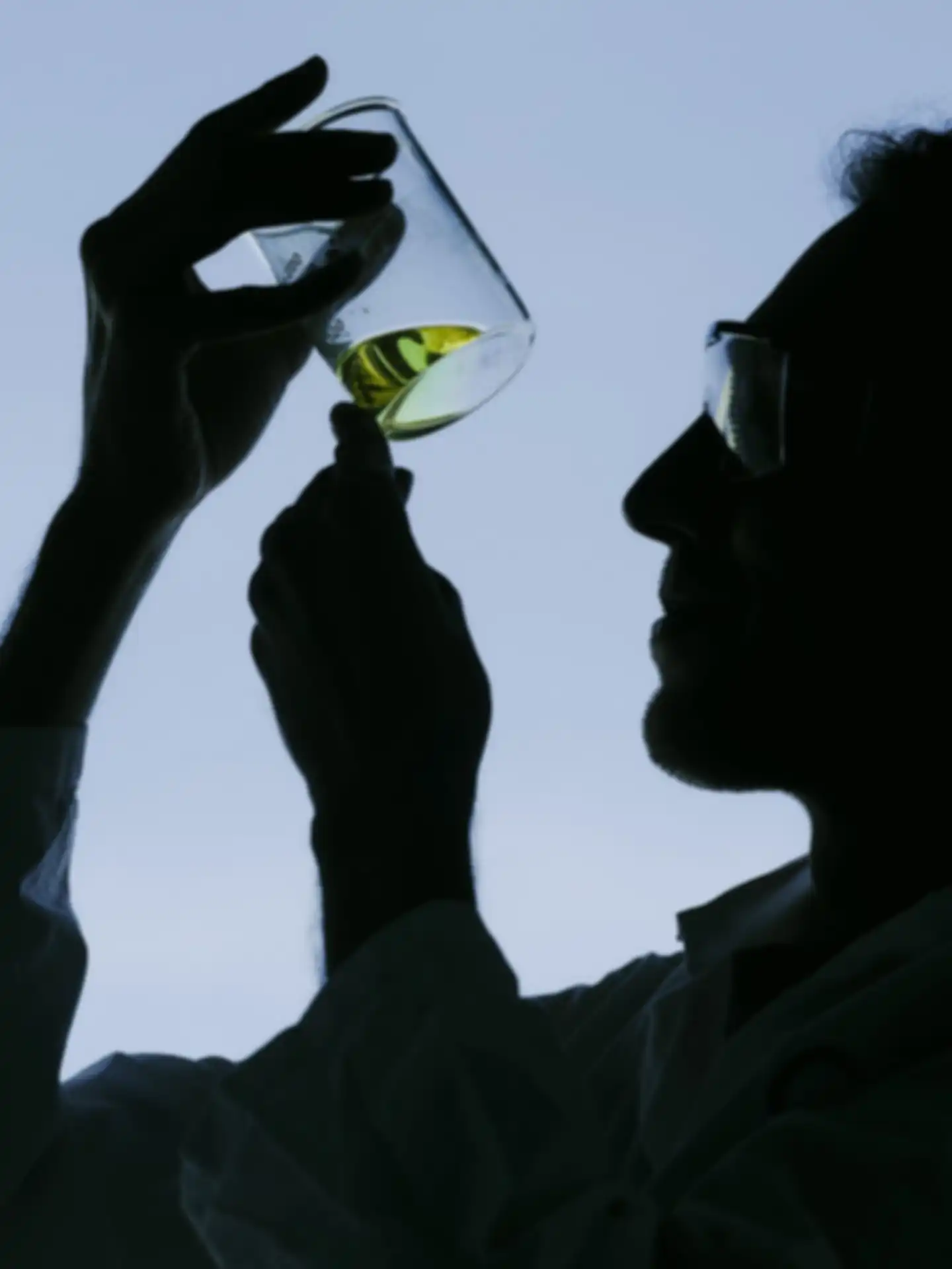 A man wearing safety goggles is holding a glass container filled with liquid in the air