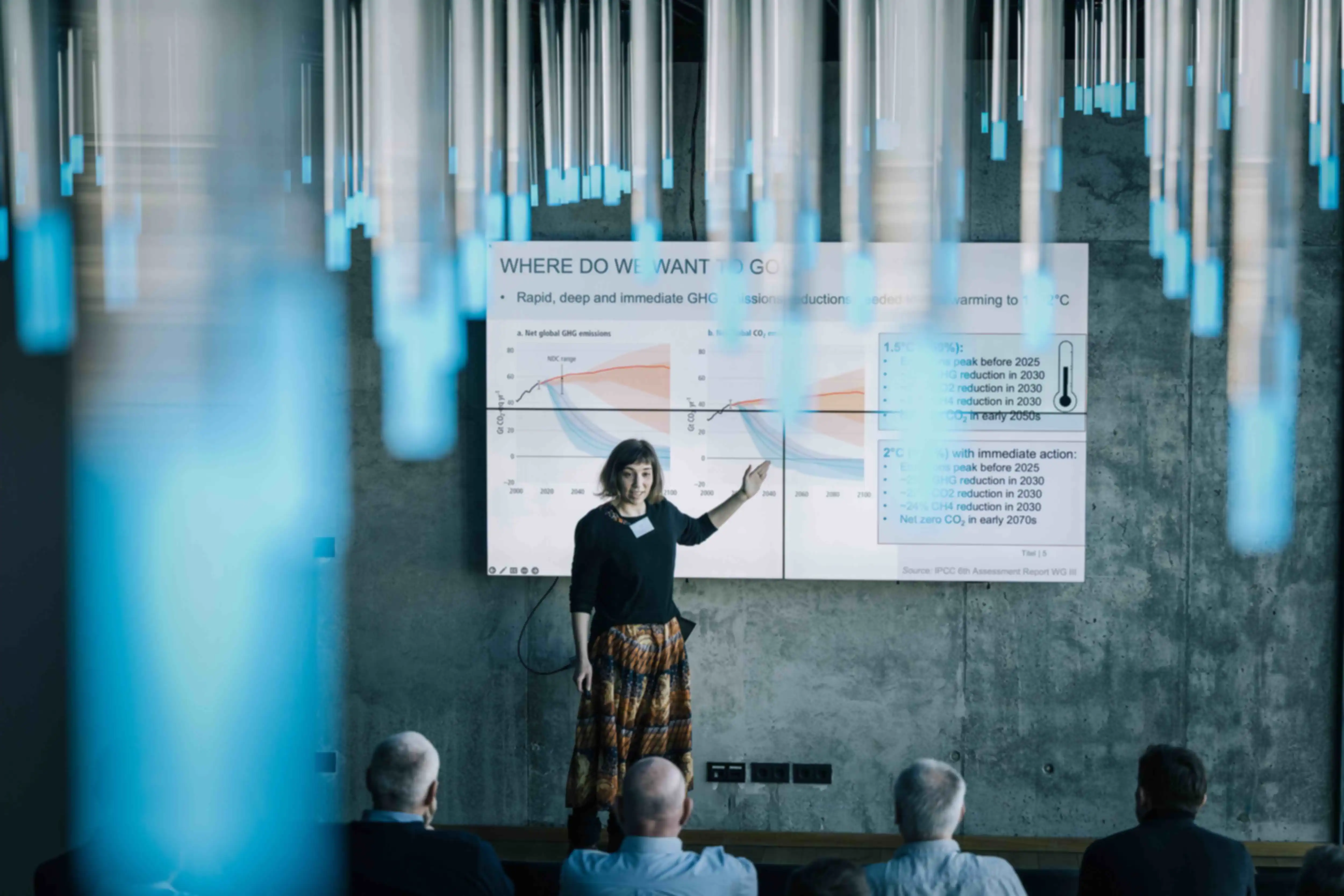 Eine Frau hält einen Vortrag, vor einem großen Bildschirm mit einer eingeblendeten Grafik