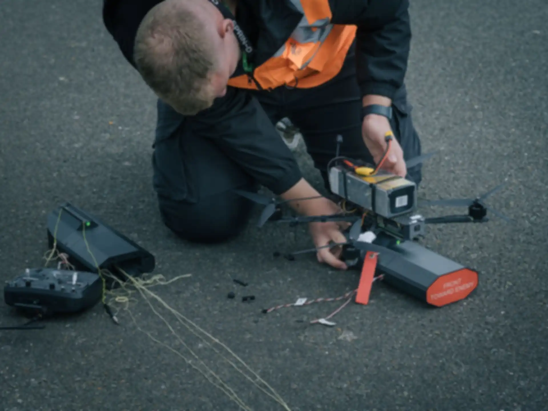 Ein Mann mit einer oragenen Veste repariert eine Drone die auf dem Boden liegt